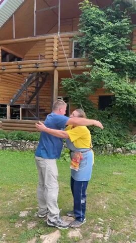 A man and a woman are standing in front of a large wooden house. The man is wearing a blue t-shirt and khaki pants. The woman is wearing a yellow t-shirt and a denim apron. They are hugging. The woman is holding a package. After a moment, they break the hug and begin to talk. The man gestures with his hands. The woman laughs.