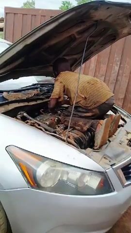 The video captures a young boy working on the engine of a silver car, likely a Honda Accord given the design of its front lights. The car's hood is fully open, propped up, revealing a dirty engine bay.

Initially, the boy is seen bent over the engine, his back to the camera, deeply immersed in the work. He is wearing a striped short-sleeved shirt and dark pants, both visibly stained with grease and dirt. He then repositions himself, squatting on the car's front fender and bumper area, with his legs inside the engine bay. From this vantage point, he continues to work on the engine with his hands. His face, now visible, shows a serious and focused expression, indicating his concentration. He is wearing sandals.

The scene is an outdoor, informal car repair area or workshop in Karshi, Nigeria. The ground is dirt, and other vehicles are parked in the background, suggesting a bustling environment. A reddish-brown structure or wall is also visible behind the car. The lighting indicates it is daytime. There is no discernible text visible in the video.