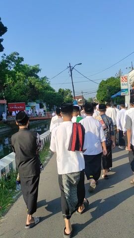 A group of men and boys walk along a street. They are wearing traditional Muslim clothing, including black peci hats. One man is wearing a black vest over a white shirt with a batik sarong. Another man has a red and black striped scarf slung over his shoulder, with a batik sarong. They are walking past a canal, with a sign for Unipdu in the background, along with an ATM for BNI. The sky is clear and blue.