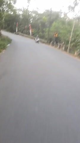 The video shows a drone's eye view of a motorcycle rider on a road. The road curves to the left and is lined with trees and foliage on both sides. There are poles with red and white flags on the side of the road. There are also some houses visible in the background. The sky is overcast.