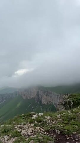 The video shows a wide, panoramic view of a mountainous landscape. The terrain is rugged, with steep cliffs and rolling green hills covered in grass. In the foreground, there are rocks and patches of grass. The middle ground features a dramatic cliff face, partially shrouded in mist and clouds. Beyond that, a series of lush green mountains recede into the distance. The sky is overcast with heavy, gray clouds, creating a moody and atmospheric feel.

The scene appears to be set in a remote, natural location. The presence of mist and clouds suggests either a high altitude or humid weather conditions. The overall impression is one of grandeur and a sense of isolation, with the vastness of the natural landscape dominating the view. There are no visible people or man-made objects, emphasizing the untouched nature of the environment. The time of day is unclear due to the overcast sky, but the soft, diffused light suggests it could be morning or late afternoon. The weather is clearly cloudy and possibly damp, adding to the dramatic and somewhat somber atmosphere. The location cues, such as the specific geological formations and vegetation, hint at a mountainous region, consistent with the provided context of Teghut, Armenia. The overall emotion conveyed is one of awe at the raw beauty of nature. There is no visible text in the video.
