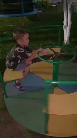 A young boy is spinning on a merry-go-round. The merry-go-round has green railings and seats that are yellow and blue. The boy is wearing a dark t-shirt with a pattern and blue shorts. He is holding onto the central steering wheel, looking at the camera with an excited expression. The background shows a grassy area with trees and other playground equipment, suggesting it is an outdoor park or playground. The lighting appears to be dim, possibly dusk or nighttime. The boy is speaking in Russian, but his words are not clearly discernible.