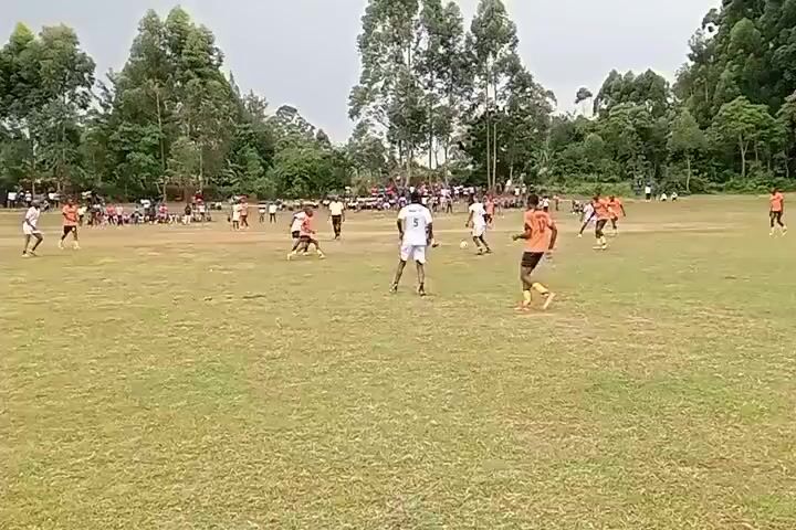 The video shows a soccer game in progress on a green grass field, surrounded by trees and a crowd of spectators along the sidelines. Players in orange and white jerseys are actively engaged in the game.  A white building with a visible soccer goal is located in the background on one side of the field. The weather appears overcast. Players are running and kicking the ball as the game continues.