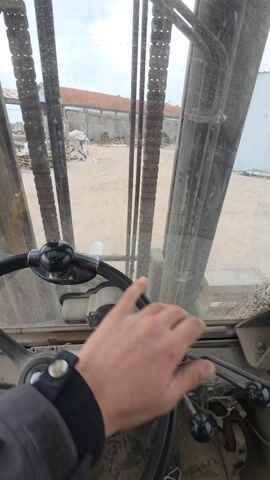 The scene is viewed from the inside of a forklift, through its windshield. A person's hand is visible on the steering wheel, and the interior of the forklift appears dusty and used. The forklift forks are visible, as well as hydraulic lines and chains used for raising and lowering the forks. Outside the forklift, a dirt yard is visible, with a building in the background with a red tile roof. Some wooden planks are stacked in the yard. The sky is overcast and grey. The forklift appears to be stationary in the video.