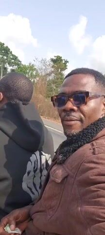 A man films himself riding on the back of a scooter, the driver wearing a black hoodie and white earbuds, with the cameraman sporting sunglasses and a brown jacket. The video is taken during the daytime on an asphalt road lined with trees. The man filming smiles for the camera as the scooter travels down the road.