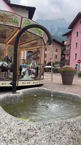 A fountain with flowing water is in the foreground, with a carousel visible in the background. The carousel has several horses, some of which are painted black and white, and others are blue and white. The carousel is decorated with lights. Behind the carousel, there are buildings with pink and brown facades, and a mountain range can be seen in the distance. There are also potted plants with red flowers. The ground is paved with cobblestones. The sky is overcast. The video shows the fountain from a medium shot, with the carousel and the town in the background. The water from the fountain is clear and splashes into the basin. The carousel appears to be stationary. There are no people visible in the video.