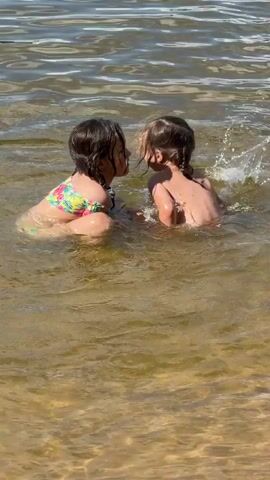Two young children are playing in a body of water, likely a lake or river, given the sandy bottom visible near the foreground. The water is calm and appears to be shallow enough for the children to stand. The sunlight suggests it is daytime, likely warm weather.

The child on the left is sitting in the water, facing the other child. She has dark hair and is wearing a colorful floral swimsuit. The child on the right is standing and facing the child on the left. She has lighter brown hair and is wearing a bikini. They appear to be interacting playfully, with the standing child reaching out to the sitting child. Both children seem happy and engaged in their activity.

The scene is set in a natural outdoor environment. The water is a murky brown-green, typical of natural bodies of water with sediment. The background shows gentle ripples on the surface of the water, indicating slight movement. The overall impression is one of carefree childhood fun in a summer setting. No visible text is present in the media file. The location is identified as Lopotovo, Russia.
