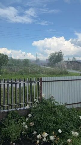 This is a vertical, medium shot video of a rural landscape on a cloudy day. The foreground features a white fence and green foliage with some white flowers. Beyond the fence, there is a grassy field with scattered trees and buildings in the distance. The sky is blue with white clouds, and there are some white specks, possibly pollen or light precipitation, visible in the air. Power lines are also visible in the upper portion of the frame. The overall impression is of a peaceful, natural setting.