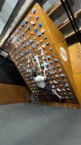 A man is bouldering on an angled plywood wall inside a building. He is wearing a white t-shirt, black shorts, and a black baseball cap. The bouldering wall is covered with numerous blue and white climbing holds, and the word “MOON” is visible on a circular logo in the upper right corner. The man moves along the holds, using his hands and feet to maintain his grip and ascend. The surrounding environment includes a concrete floor, corrugated metal roofing, and wooden walls and structural elements.