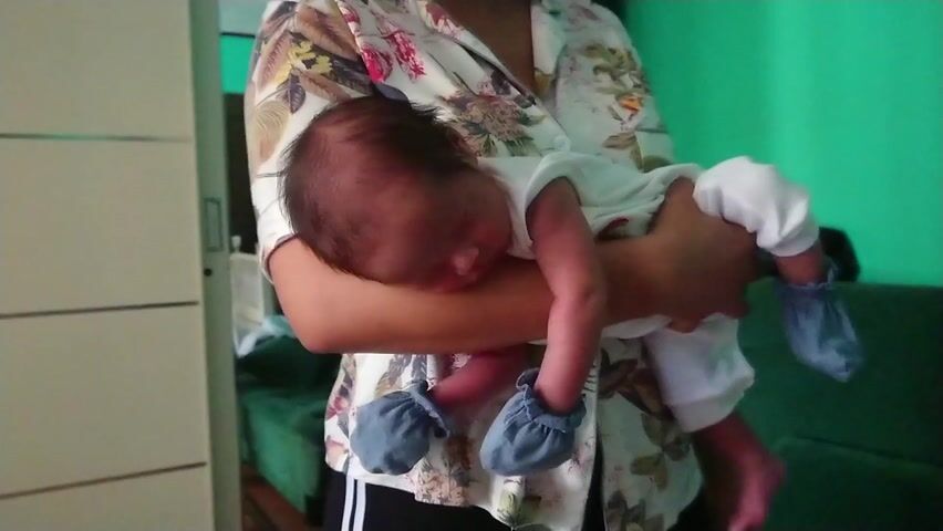 In a home setting, an adult, partially visible and wearing a floral-patterned button-up shirt, gently holds a sleeping newborn baby. The baby, with dark hair, is dressed in a white onesie and light blue foot coverings. The adult's arms are cradling the baby, who appears to be soundly asleep, lying on its stomach across the adult's arm. In the background, a light-colored wardrobe or cabinet is visible on the left, and a bright green wall with a darker furniture piece is on the right. The scene conveys a sense of quiet and tenderness. No text is visible in the frame. The video lacks specific visual cues to confirm the provided location context of Coubron, France.