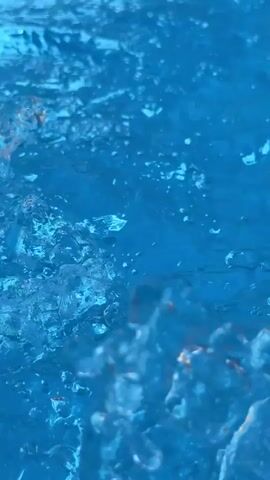 This video showcases a close-up view of water flowing into a swimming pool. The water is a vibrant blue, reflecting the sunlight. Bubbles and ripples are visible on the surface, indicating movement and aeration. The camera focuses on the waterfall effect as the water cascades from a tiled edge into the pool. The tiles are a mosaic of various shades of blue, creating an attractive border.

In the background, glimpses of lush green foliage and a glimpse of what appears to be palm trees suggest a warm, tropical or Mediterranean climate. The bright lighting indicates it is daytime, likely sunny. The scene evokes a sense of relaxation and tranquility, typical of a pool environment. The sound of the cascading water is likely a prominent element, further enhancing the serene atmosphere. The location is identified as Seixal, Portugal, which aligns with the visual cues of sunshine and greenery. There is no visible text in the media file.