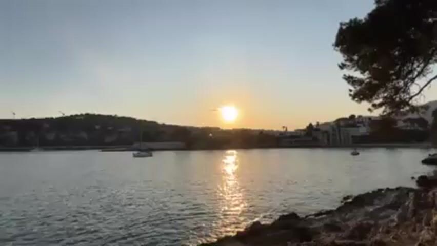 Here is a description of the video:

The video begins with a scenic view of a calm sea as the sun sets in the distance. The sun's reflection creates a shimmering line across the water, and several sailboats are visible in the harbor. The shore is lined with rocky terrain, and some buildings can be seen further inland amidst the trees. The scene is peaceful and bathed in the warm light of the setting sun.

The camera then flips to show a young couple. The man and woman are both wearing sunglasses and looking directly at the camera, which is positioned above them.