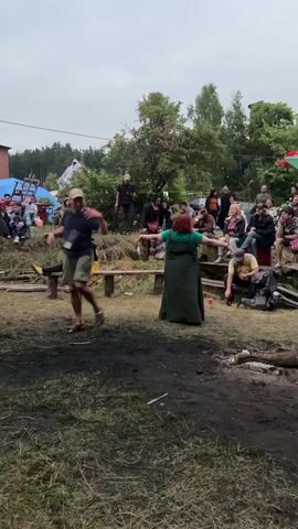 The video shows an outdoor gathering with several people. In the foreground, two people are dancing energetically. One is a man in a dark t-shirt and shorts, and the other is a woman in a long green dress. Behind them, a group of people are sitting on a wooden bench and on the ground, watching the dancers. Further in the background, there's a stage with musicians performing under a white canopy illuminated by purple and green lights. More people are visible in the background, some sitting and some standing. The ground is covered with grass and dirt, with some scattered logs and debris. The overall atmosphere appears to be festive and casual.