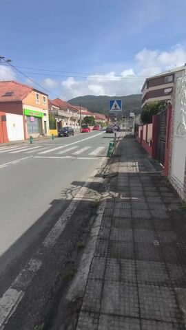 The video depicts a quiet street scene in a small town. A pedestrian crossing is visible with a sign. Cars are parked on the side of the road and some are driving in the distance. Houses line both sides of the street, and a mountain is visible in the background. The weather appears sunny and bright, casting shadows on the sidewalk.