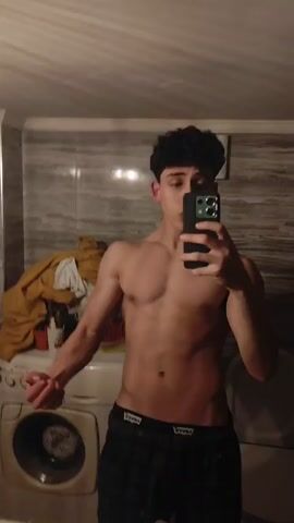 A young man with curly hair is taking a selfie in a bathroom mirror. He is shirtless and wearing dark-colored shorts with a logo waistband. The bathroom has marble-patterned walls and contains a washing machine to the left and a toilet to the right. The man flexes his arm and poses for the camera. The lighting is dim.