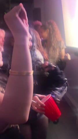 The video shows what appears to be a group of people at a social event or party. The lighting is low, creating a club-like atmosphere. The people are interacting with each other, laughing, and moving around. Several people are holding red plastic cups, suggesting they are drinking. The video is shaky, likely due to someone recording while moving. One person is briefly seen near a laptop and a colorful light source. The general mood seems festive.