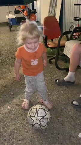 In this short video clip, a young child with blonde, messy hair is playing with a soccer ball. The child is wearing an orange t-shirt with a white bunny design and grey camouflage-patterned pants. They are barefoot. The child is kicking the soccer ball.

The setting appears to be an outdoor, rural area, likely a backyard or yard. There are several objects visible in the background, including a tiller or small tractor, a red chair on wheels, a bicycle, and what looks like corrugated metal fencing. The ground is covered in gravel and dirt.

The lighting suggests it is either late evening or early morning, as the light is dim and there are hints of darkness. The overall mood of the clip is playful and energetic, with the child fully engaged in their activity. There is no visible text in the media file. The location context provided is Әуезов ауылдық округі, Kazakhstan.
