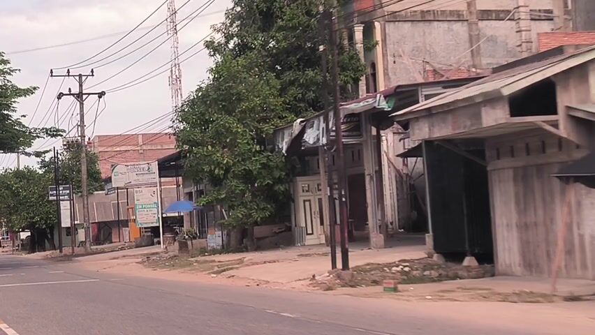 The video presents two brief street-level perspectives captured from a moving vehicle in Matangpineung, Indonesia. Both clips depict a paved road lined with buildings, utility poles, and trees under an overcast sky.

The first segment (0:00-0:02) showcases a street with a mix of single and multi-story structures, likely containing shops and residences. Numerous power lines are visible overhead, supported by wooden utility poles. Lush green trees flank the road, and a tall communication tower stands prominently in the background. Several signs are visible, displaying text such as "Pelukis," "BSI" (Bank Syariah Indonesia), "ZIR PONSEL," and "OPPO." A small blue umbrella is positioned in front of one of the establishments.

The second segment (0:02-0:04) continues down a similar street. Two people, both wearing headscarves, are seen riding away from the camera on a motorcycle, a common mode of transport. Other vehicles, including cars and motorcycles, are present in the distance. The buildings along this stretch also feature tiled roofs and open shopfronts. A small yellow warning sign is visible on the roadside. In the far background, the dome of a mosque can be faintly discerned.

The time of day is daytime, with the overcast conditions providing soft, diffused lighting. The scene portrays typical daily life and infrastructure within a town or village setting in Matangpineung, Indonesia.