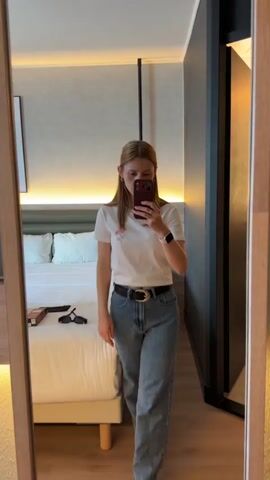 A young woman with blonde hair pulled back stands in front of a mirror, taking a selfie with her phone. She is wearing a white t-shirt with a small pink flower design, blue jeans, a dark belt with a silver buckle, and a watch on her left wrist. Her hair is parted in the middle and falls to her shoulders. She has light makeup on, including lipstick. The background appears to be a hotel room. There is a bed with pillows behind her and a framed picture on the wall to her left. To her right, a dark wood door is partially visible. A bedside table with some items on it is also visible in the reflection. She is holding a burgundy phone with a three-camera system. The lighting in the room is warm.