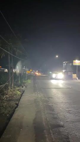 The video shows a nighttime street scene with a paved road and a sidewalk. Several motorcycles are visible on the road, with their headlights and taillights illuminated. The road is lined with trees and utility poles. Buildings and signs are visible on the right side of the road. The overall atmosphere is dark and somewhat hazy, suggesting the presence of streetlights and possibly some atmospheric moisture. The video captures the movement of the motorcycles as they travel down the road, creating streaks of light due to the long exposure.