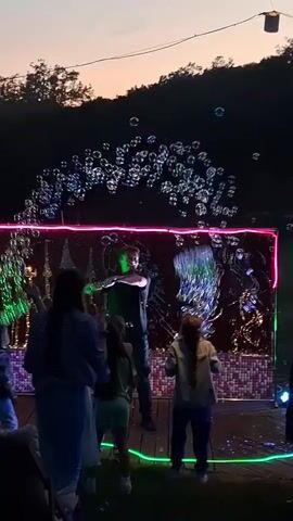 In a video taken outdoors at dusk, a man operates a bubble machine for a crowd of mostly young children. The machine is situated in front of a decorated wall with a pink neon bar across the top and a green bar at the base. The man is facing away from the camera, but his dark hair is visible. The children are facing the machine, reaching up to catch the bubbles. The background includes trees and a dusky sky. A small spotlight illuminates part of the scene from the right.