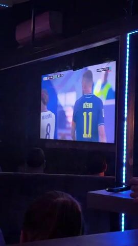A large television screen displays a soccer match. A player wearing a blue jersey with the number 11 and the name "DZEKO" on the back is the primary focus on the screen. To the left of the screen, another player in a red and white striped jersey with the number 8 is visible. The score displayed on the screen indicates a game between two teams, with one team having a score of 0 and the other also 0. The time on the game clock reads 42:26. The broadcast channel is identified as ARENASPORT. The television is mounted on a wall in a dark room, with people seated in front of it. The lower portion of the screen shows the top of the heads of two individuals watching the game. There are blue LED lights visible on the right side of the screen. The overall environment appears to be a viewing area, possibly a sports bar or lounge.