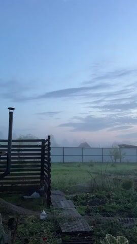 The media file captures a serene outdoor scene at dusk in Emel'yanovo, Russia. The primary subject is a rural backyard with a fence, garden, and a wooden structure. The sky is a prominent feature, displaying a gradient of soft blues and pinks, indicating the transition from day to night. Wispy clouds are scattered across the sky, adding texture to the twilight atmosphere.

In the foreground, a wooden fence forms a boundary on the left side of the frame, with a chimney-like structure attached to it. A small wooden pathway leads into the garden area, which is filled with lush green plants and some uncultivated vegetation. In the mid-ground, a white metal fence stretches horizontally across the scene, separating the backyard from what appears to be a neighboring property or open field. Beyond the white fence, the silhouettes of houses are faintly visible through a light mist or fog, adding a sense of depth and mystery. The overall setting suggests a quiet, rural environment.

The time of day is clearly evening, given the dusky sky and the low light conditions. The presence of mist hints at cool, humid weather, typical of early mornings or evenings in certain climates. There are no people or animals visible in the frame. The mood conveyed is peaceful and tranquil, with a touch of melancholic beauty due to the fading light and mist. No text is visible in the media file.