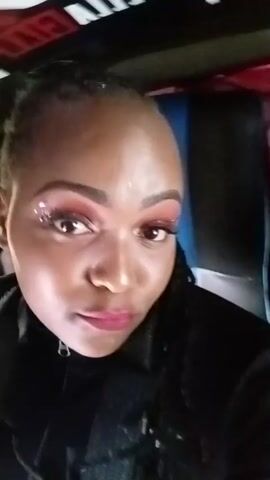 A close-up shot features a dark-skinned woman, wearing a black jacket, looking directly at the camera. Her makeup is noticeable, with prominent red eyeshadow and light pink lipstick. She smiles and poses subtly throughout the video. The background consists of dark, undefined car interior elements. Loud, rhythmic music plays throughout the video, creating a party-like atmosphere.