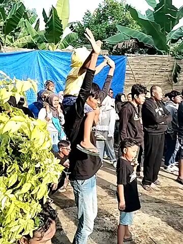 The video depicts a crowd of people gathered outdoors. In the foreground, a man carries a child on his shoulders, both raising their hands. Many onlookers are visible, including women wearing hijabs. A blue tarp hangs in the background, alongside tropical plants. The camera then pans to a larger group of people watching performers dressed in black attire. They are accompanied by music and appear to be engaged in a traditional dance or performance. The environment seems to be a rural area with trees and simple structures in the background. Many people in the crowd are recording the event with their phones.