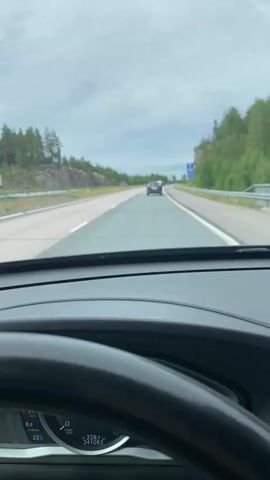 This media file presents a point-of-view shot from inside a car, looking forward through the windshield. The driver's side dashboard and steering wheel are visible in the foreground, with a digital display showing speed (0.8 km/h), temperature (23°C), and a trip odometer (229.8 km, 241082). A clock indicates 16:14.

The scene is a highway in a natural setting, with lush green trees and rocky outcrops lining both sides of the road. The sky is overcast with clouds, suggesting a temperate climate. The road itself is paved and appears to be a multi-lane highway, indicated by the white dividing lines. A dark-colored car is visible in the distance, traveling in the same direction as the camera car. A blue road sign with white text and symbols is visible on the right side of the road.

The overall impression is one of a relaxed journey on a highway, likely in a rural or semi-rural area. The lighting suggests it is late afternoon. The location context places this drive in Porvoo, Finland, a region known for its picturesque landscapes.