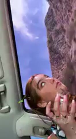 The video shows a woman inside a vehicle, likely a car, with the roof and a sun visor visible. The background outside the car features a clear blue sky with some white clouds and a large, rocky, brown cliff or mountain formation, consistent with a desert or mountainous region like Desert Hot Springs, United States.

The woman has brown hair and is seen looking upwards and away from the camera in the first frame, with her mouth slightly open, appearing to react to something or speaking. She is holding a small spray bottle or container, possibly applying makeup or perfume. In the second frame, she turns to look at the camera, making a peace sign or "V" gesture with her hand. The scene takes place during the daytime under clear weather conditions. Audio includes sounds and speech, with someone saying "Oh" and "Not that. Yeah, I did." No visible text is present in the video.