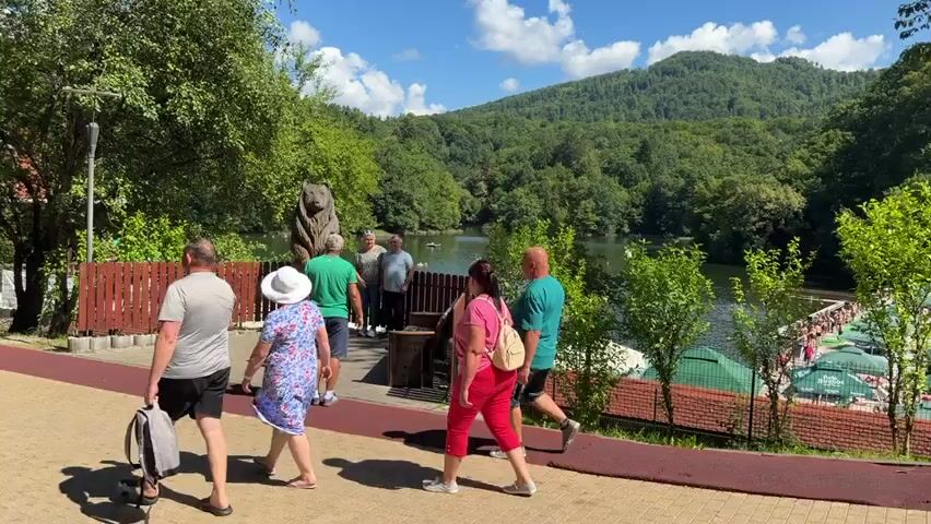 This is a daytime, outdoor, medium shot video with a slight downward angle. The scene is a paved walkway next to a wooden fence and a body of water. A group of people are walking on the path, while others are standing near the water. To the left, a man in a gray t-shirt and black shorts is walking away from the camera, pulling a wheeled bag. A woman in a floral dress and a white hat is walking ahead of him. Behind them, another woman in a pink shirt and red pants is walking towards the camera. To the right of the path, a wooden sculpture of a bear stands next to a fence. Several people are gathered near the bear sculpture, with two men posing for a photo. Further to the right, there is a body of water with a forested hill in the background. Several green umbrellas are visible on the far side of the water, suggesting a recreational area. The sky is blue with scattered white clouds.