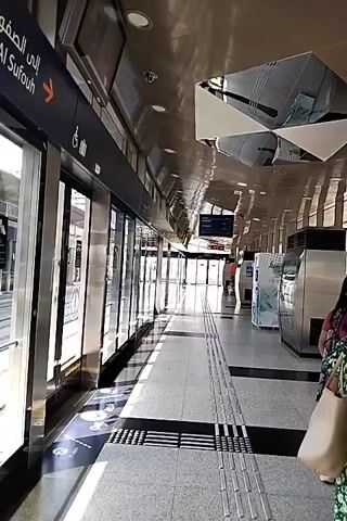 The video shows a deserted train platform with doors closed. The platform is modern and clean, with metallic walls and a tiled floor. There are signs in Arabic and English indicating the direction of travel. A few people can be seen in the background, waiting for the train. The lighting is bright, suggesting daytime. The overall atmosphere is one of anticipation and efficiency, typical of a public transportation hub.