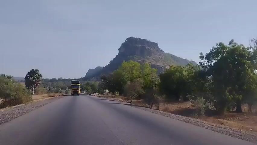 The video shows a vehicle driving along a black asphalt road. The road has gravel shoulders on either side. In the distance, a large, rocky hill is visible. There are scattered trees and bushes along the sides of the road. A large, yellow and black truck is driving towards the camera on the road. The sky is light blue and clear.