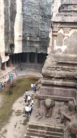 The video shows a group of people exploring a large, ancient rock-cut structure. The environment appears to be outdoors with rough, textured rock walls forming a cavernous space. The ground is uneven and wet in places. Several individuals are walking and observing the architecture, which features carved pillars and intricate details. A prominent element in the foreground is a large, elaborately carved stone structure with an elephant carving visible. The video focuses on a few individuals in wheelchairs, being assisted by others, as they navigate the complex. Some individuals are dressed in traditional Indian clothing like sarees, while others wear casual attire. The scene conveys a sense of historical exploration and accessibility being provided to visitors.