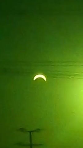 The video shows a crescent-shaped light in the sky, which appears to be the sun during a solar eclipse. The sky is green, and there are some indistinct lines or objects visible in the upper and lower portions of the frame, possibly power lines or tree branches. The light source is stationary throughout the short clip.