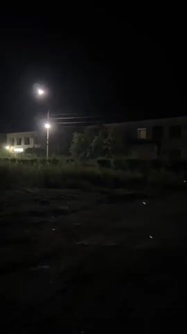 The video shows a dimly lit outdoor scene at night. The camera is moving slowly. In the foreground, there is dark, uneven ground, possibly dirt or gravel. In the mid-ground, there is tall, overgrown grass and vegetation. Behind the vegetation, a long, light-colored building with several windows is visible. A street lamp with a bright light is illuminating the area. There are power lines running horizontally across the scene. In the distance, there are more buildings and trees. The overall atmosphere is dark and somewhat eerie. There are a few faint green lights that appear to be reflections or possibly insects. No people are visible in the video. The camera movement suggests the person recording is walking or slowly driving through the scene.