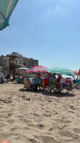 A sunny day at a crowded beach. The camera is low to the sand, looking towards the ocean. In the foreground, two sets of bare feet are visible. Behind them, several beach umbrellas are set up, providing shade for people sitting on chairs. There are multiple beach chairs, some with colorful towels draped over them. In the middle ground, a lifeguard stand is visible on the left. In the background, buildings line the coast, and people are visible in the water. The sky is clear and blue.