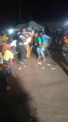 The video shows an outdoor nighttime scene at what appears to be a celebration or event. A group of people are gathered, with some individuals dancing in the center of the frame. Several bills are scattered on the ground in front of the dancers. The lighting suggests artificial illumination, likely from lights set up for the event and possibly from flashlights. In the background, there is a tent structure and a crowd of onlookers. The overall atmosphere seems lively and festive.