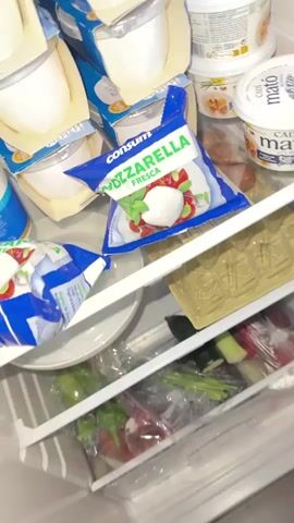 A hand reaches into a refrigerator and picks up a package of mozzarella. The refrigerator is filled with various dairy products, including yogurt and cheese. The mozzarella package is blue and white with an image of mozzarella balls and tomatoes. Another hand is visible in the lower left corner of the frame. The video ends with the hand holding a block of cheese.
