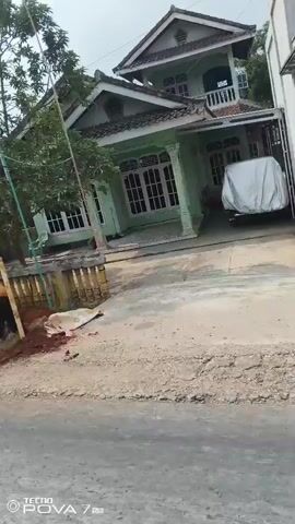 The video shows a residential street with a house in the background. In the foreground, two individuals are engaged in digging work. One person, wearing a yellow shirt and dark pants, is actively digging into the reddish-brown soil with a tool. Another person, in a green shirt and dark pants, is also working on the ground nearby. A white tarp or sheet is visible on the ground between the two workers. A car covered with a white tarp is parked under a carport attached to the house. The house itself appears to be a two-story structure with white walls and a brown tiled roof. A fence with decorative columns runs along the front of the property. The environment suggests an outdoor setting, possibly a suburban or rural area, during daylight hours.