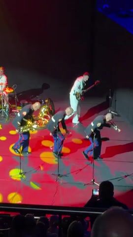 A live musical performance takes place on a stage illuminated by red and yellow lighting. Five musicians are visible. In the foreground, three individuals in military dress uniforms with blue pants and red jackets are playing instruments. The leftmost individual plays a saxophone. The middle individual plays a tuba. The rightmost individual plays a violin. Behind them, a person in a light-colored suit plays a bass guitar. To the far left, another person in a similar light-colored suit plays drums. The audience is visible in silhouette in the bottom of the frame.