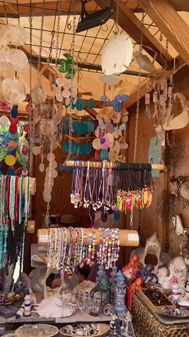 This is a video of a market stall filled with various handmade jewelry and decorations. The stall appears to be outdoors, possibly under a canopy, as indicated by the exposed wooden beams and fabric ceiling. The items displayed include necklaces, bracelets, earrings, and wind chimes, many of which are made from shells, beads, and colorful stones. The merchandise is hung from metal chains and wooden racks, creating a vibrant and eclectic display. The overall atmosphere suggests a beachside or craft market setting. There are no people visible in the frame.