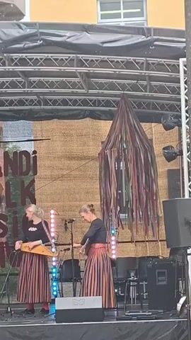 A folk music festival is underway on an outdoor stage. Four women are performing, two playing string instruments, one playing a dulcimer, and one playing a double bass. The backdrop features a banner that reads "Viljandi Folk Music Festival". Two of the women are wearing traditional striped skirts, and all are wearing black tops. A decorative, ribbon-draped structure hangs from the stage rigging. The lighting is dim, with stage lights illuminating the performers.