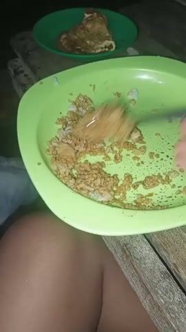 The video shows a close-up of a person's leg and arm as they eat from a light green bowl filled with rice and what appears to be fried meat or vegetables. The person is using a spoon to mix and eat the food. In the background, a green plate with a piece of cooked food is visible, along with a wooden surface. The lighting is dim, suggesting it is nighttime.