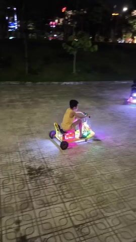 At night, two boys are riding light-up toy cars on a tiled plaza in a park. The plaza has a patterned tile surface. The first boy, wearing a yellow shirt, rides a multicolored toy car with red, orange, and white lights. The second boy, wearing a dark shirt, rides a toy car with purple lights. The background features trees, grass, and lights from buildings in the distance. The plaza is surrounded by low stone walls with grass behind them. The video follows the boys as they ride around the plaza.