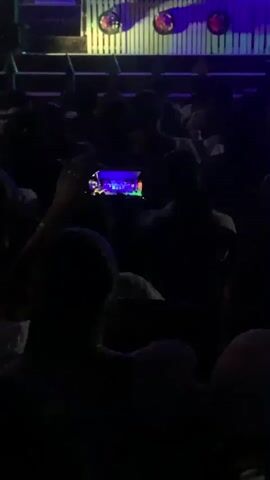 The video shows a dimly lit indoor venue, likely a theater or concert hall, with an audience seated in rows facing a stage. The stage is illuminated with bright blue and purple lights, and a group of people are visible on stage, some appearing to be performers. One audience member in the foreground is holding up a smartphone to record the event, with the phone's screen displaying a colorful image of the stage. The atmosphere appears to be that of a live performance, with the audience immersed in the experience.