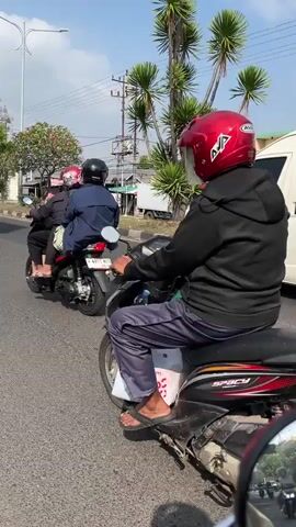This media file captures a street scene in Ngaban, East Java. The primary subjects are two individuals riding a scooter, with the person in the foreground wearing a black hoodie and a red helmet. They are traveling on a paved road alongside other vehicles, including a white van and a truck. The setting appears to be a moderately busy street with buildings, trees, and power lines visible in the background. The time of day seems to be daytime, indicated by the bright sunlight and clear blue sky. The weather appears to be fair and sunny.

The activity depicted is simply transportation; the individuals are riding a scooter on a public road. There are no strong emotional cues visible as the subjects are primarily seen from behind. Notable details include the license plate of the scooter, which reads "W 5943 WBL 05-26". The presence of tropical trees and the style of buildings suggest a warm climate. The road is asphalt, and there are markings indicating lanes. The overall impression is a typical day on a street in a populated area in Indonesia.