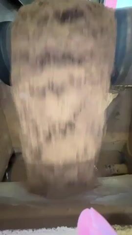 The video shows a close-up, shaky shot of a substance, possibly dirt or soil, being poured from a large cylindrical object into a box or container. The substance is brown and granular, and it appears to be falling in a continuous stream. A pink object, possibly a finger, is visible in the lower right corner of the frame, suggesting a human presence. The background is blurred and indistinct, but appears to be an indoor environment. The lighting is dim, and the overall impression is one of a somewhat messy and industrial process.