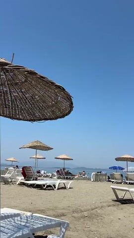 The video shows a sandy beach with several umbrellas and lounge chairs. People are seen relaxing on the chairs and walking around. The ocean is visible in the background, and the sky is clear and blue.